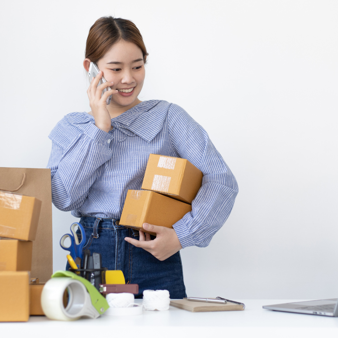 A woman holding parcels and talking on the phone, with packing materials and a laptop on the desk nearby.