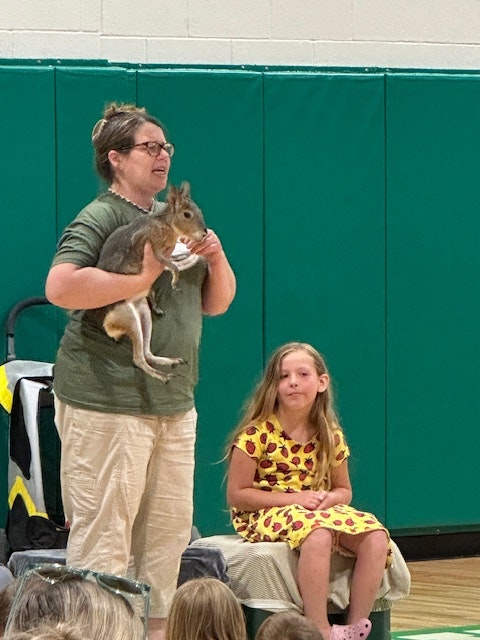A person holds a kangaroo in front of an audience, with a child sitting nearby in a yellow dress with red patterns.