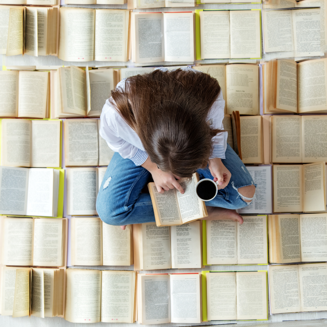 A person seated amidst many open books, engrossed in reading one, while holding a cup of coffee.
