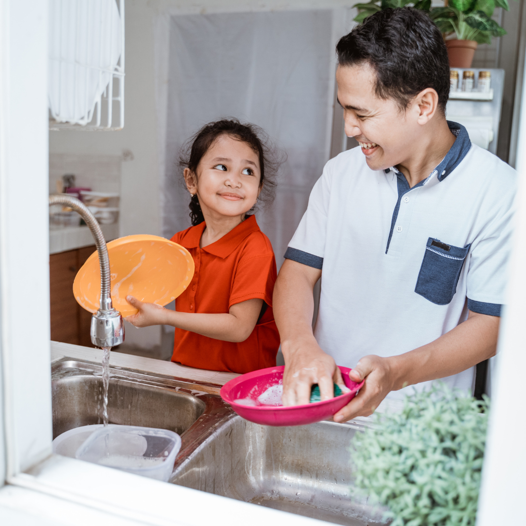 A child and an adult smiling while washing dishes at a kitchen sink.