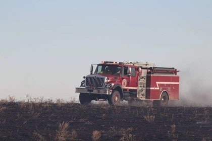 A fire truck is driving across a burnt field with smoke in the background.