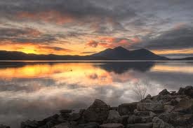 A tranquil lake reflecting a vibrant sunset, surrounded by rocky shores, with silhouetted mountains in the background.