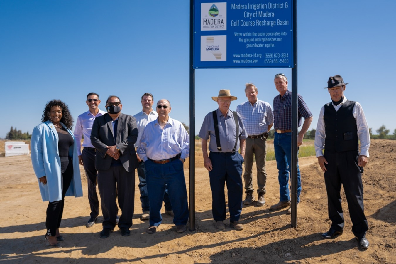 A group of people standing next to a sign for "Madera Irrigation District & City of Madera Golf Course Recharge Basin" under a clear blue sky.