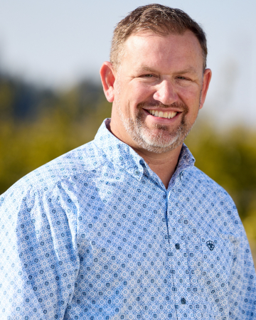 Smiling man in a patterned button-up shirt outdoors with a blurred background.