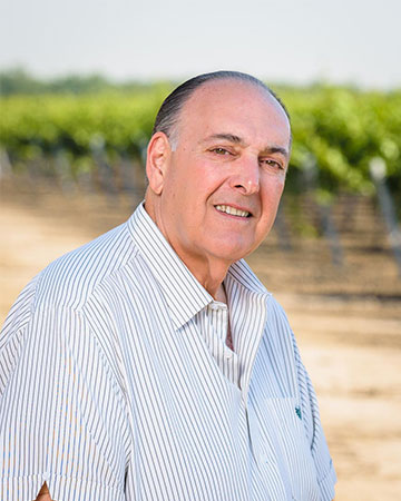 A person smiling, standing in front of a vineyard, wearing a striped shirt.