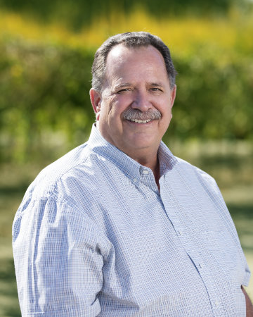 A person smiling outdoors, wearing a light-colored shirt, with greenery in the background.