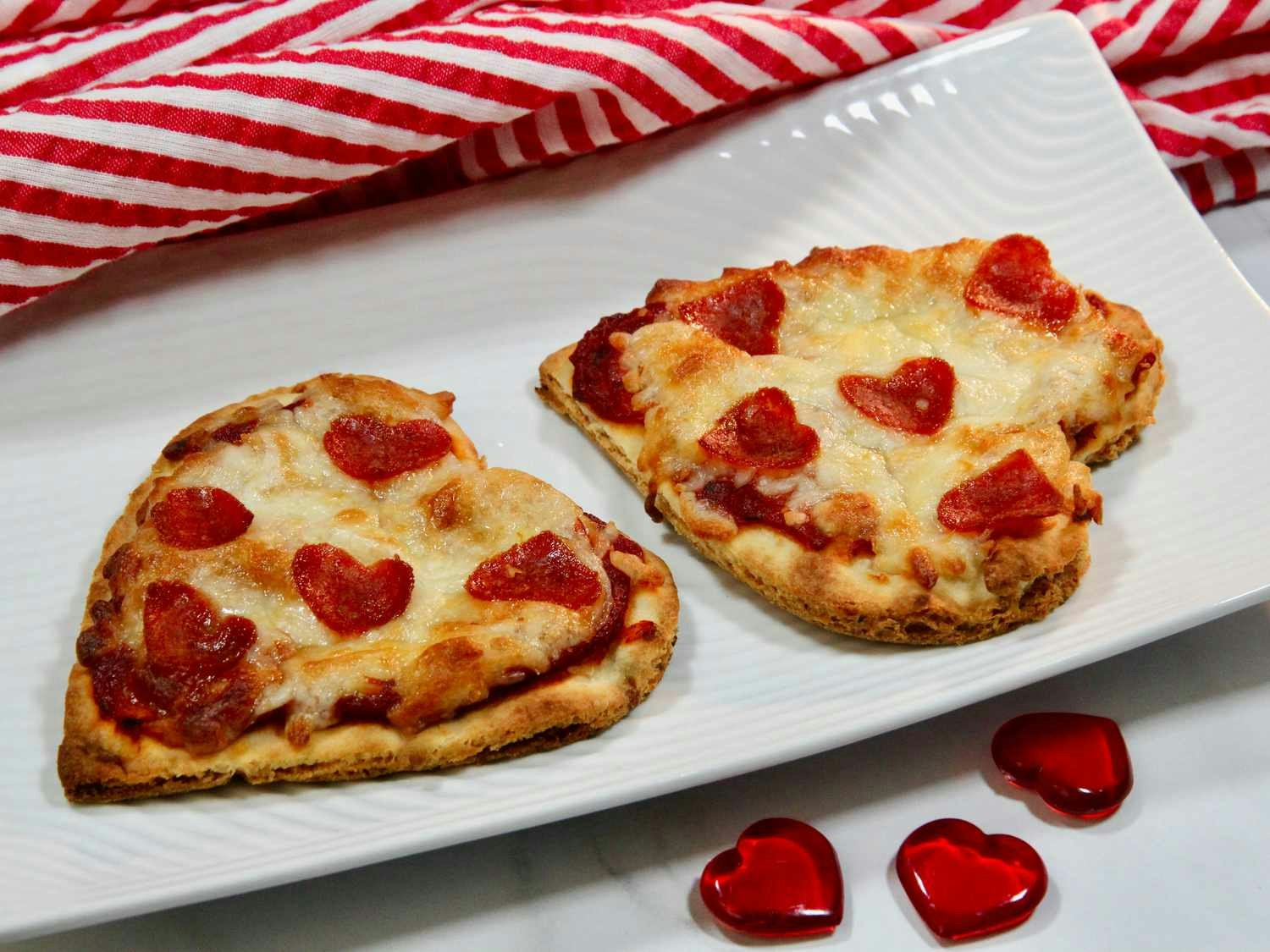 Heart-shaped pizzas with heart pepperonis on a white plate, red striped cloth, and heart-shaped decorations.