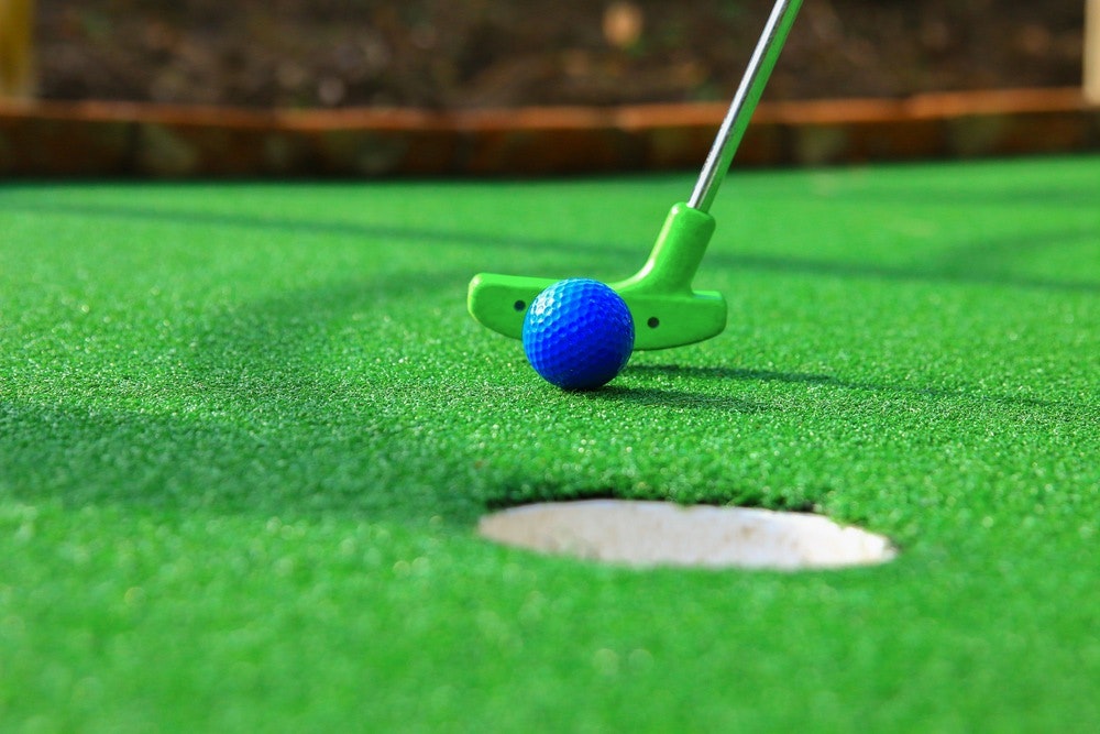 Mini-golf putting, blue ball, green club, artificial turf, hole-in-one attempt.