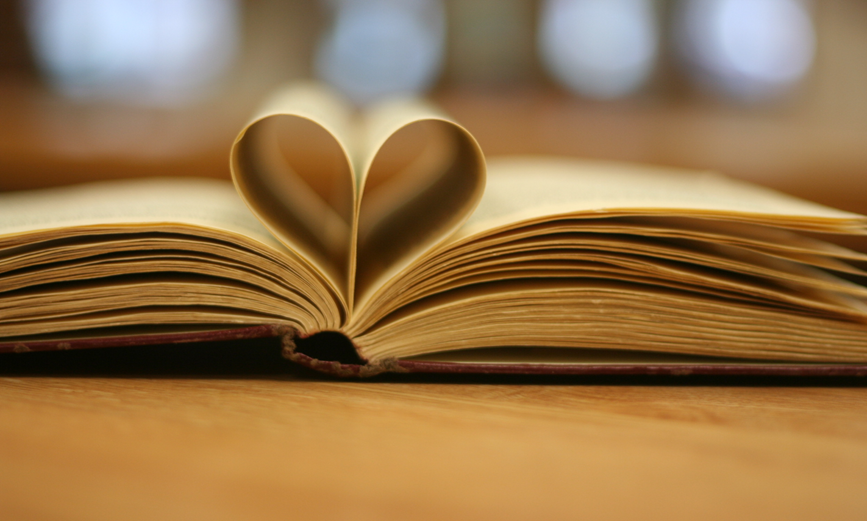 An open book with its pages forming a heart shape, resting on a wooden surface. The background is softly blurred.