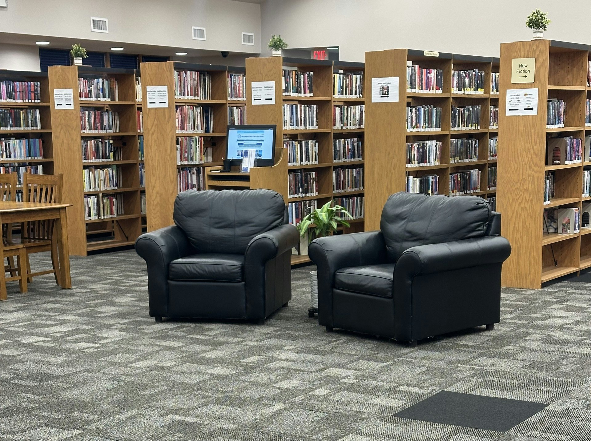 The image shows a library interior with two black chairs, wooden bookshelves filled with books, and a computer station.