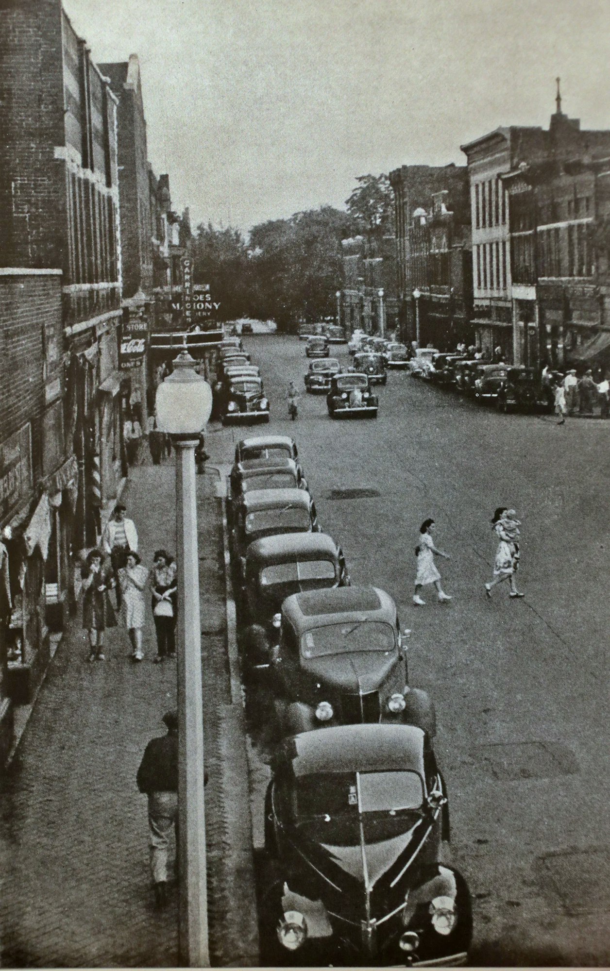 Vintage street scene with parked cars, people walking and crossing the street, and old buildings lining both sides.