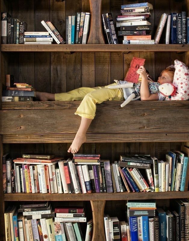 A child lies on a bookshelf, reading with a toy, surrounded by rows of books in a cozy setup.
