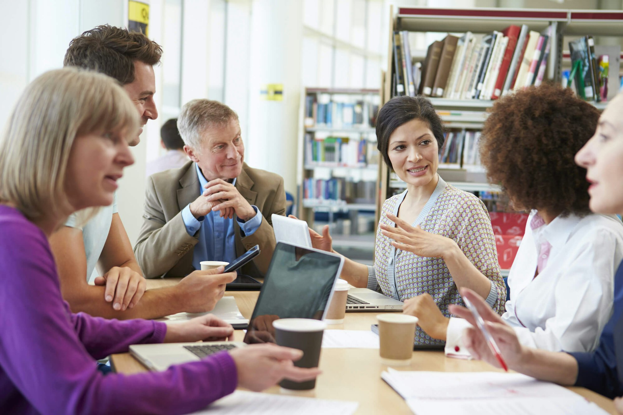 Five professionals in a meeting with digital devices and papers, engaged in discussion.