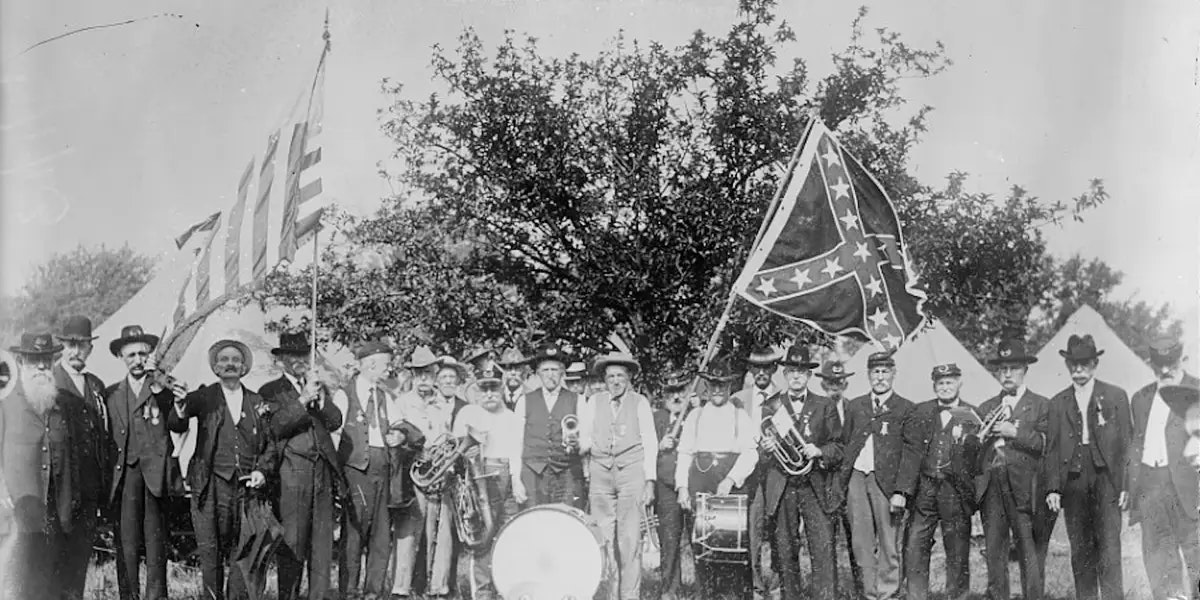 A group of people poses outdoors with musical instruments, holding American and Confederate flags, in a historical setting.