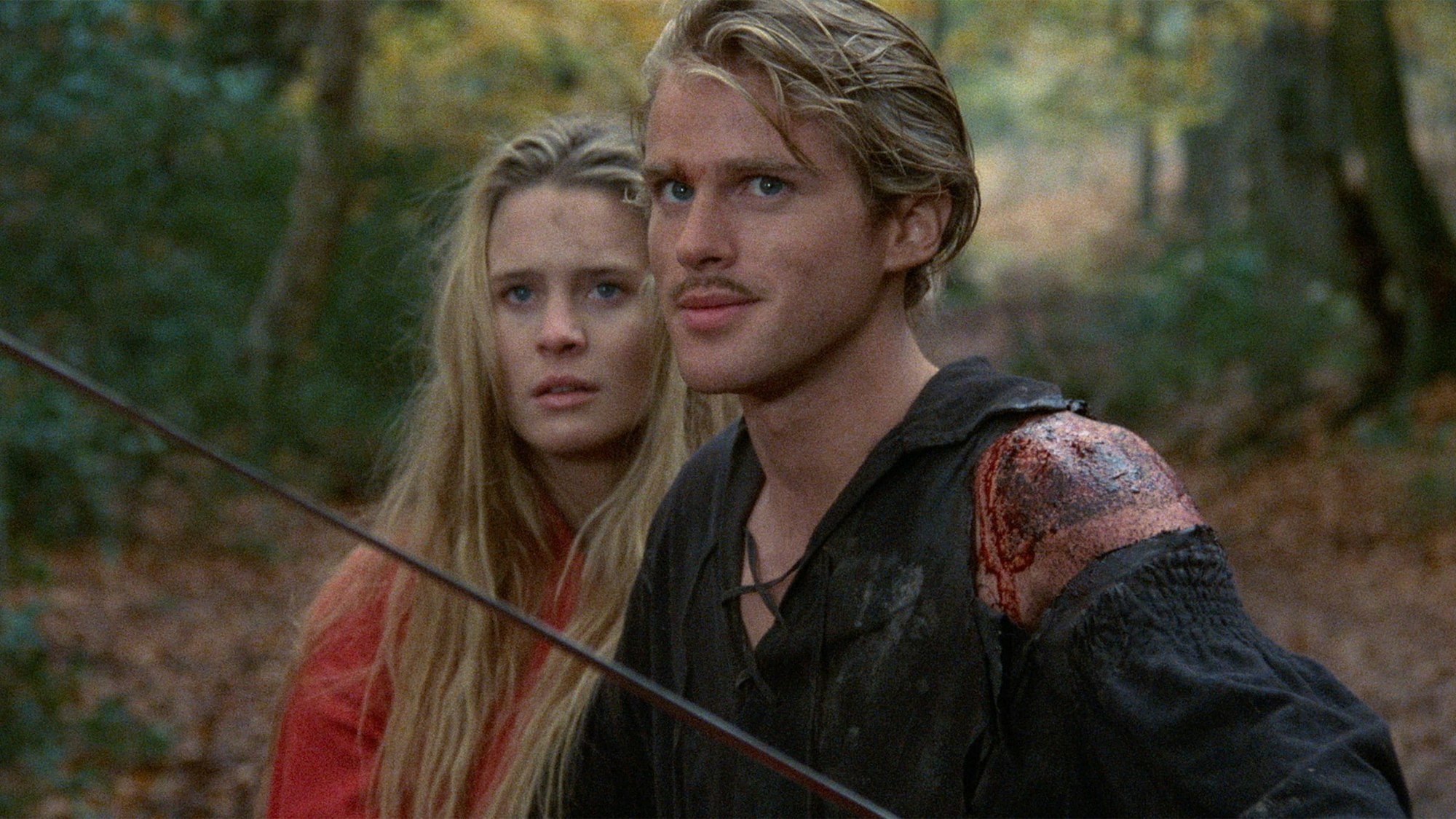 A man with a wound on his shoulder stands beside a woman in a forest setting, looking tense.