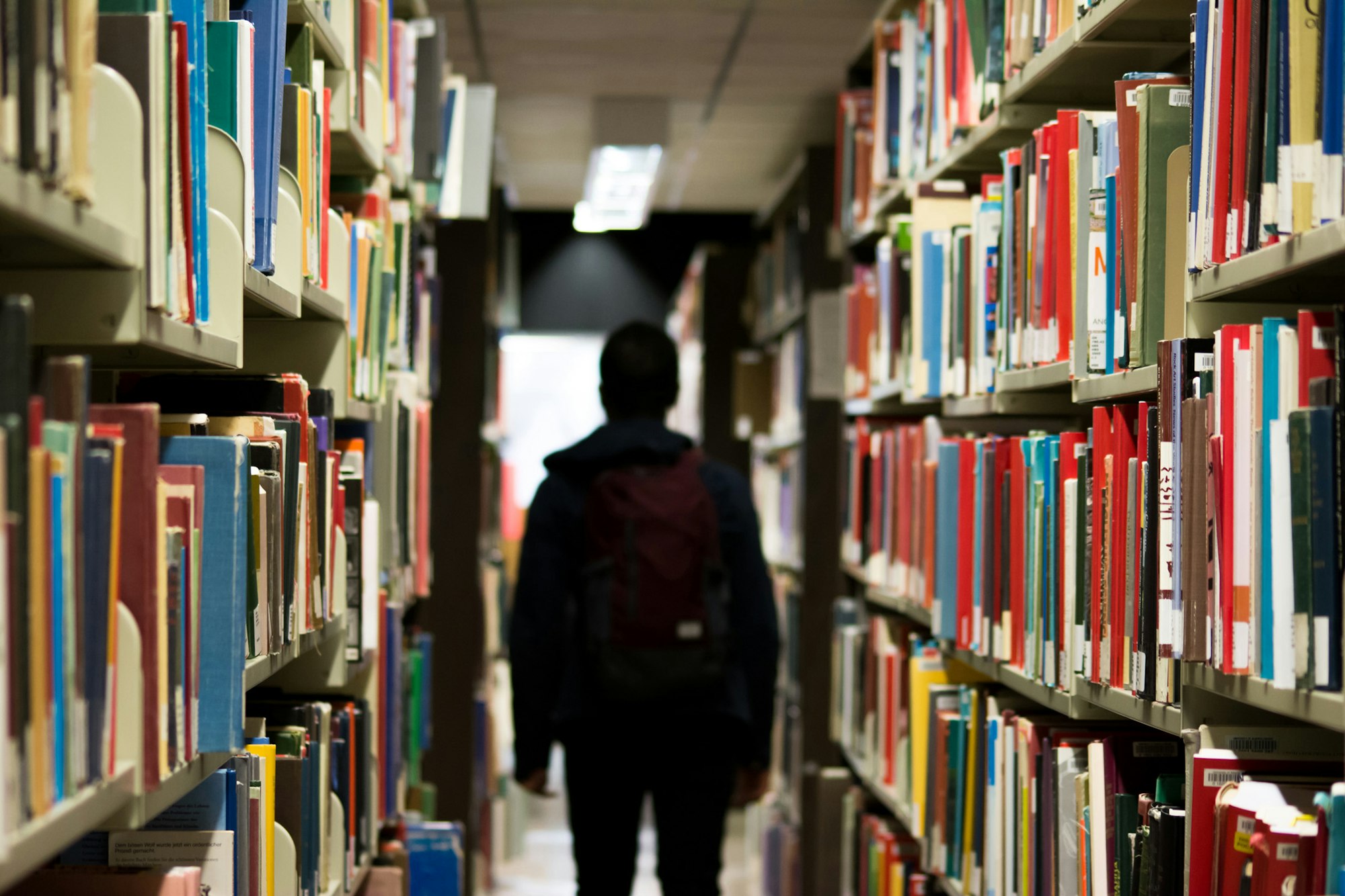 Person with backpack standing between rows of books in a library.