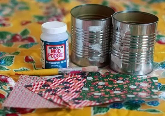 Craft supplies: 2 cans, Mod Podge, brush, and patterned paper on a colorful tablecloth.