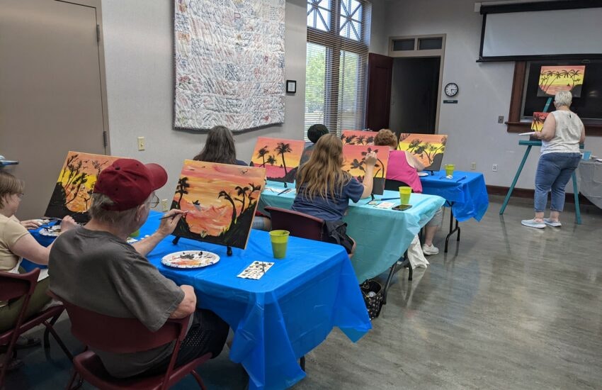 A group of people participate in a painting class, creating tropical sunset scenes on canvases at a community center.