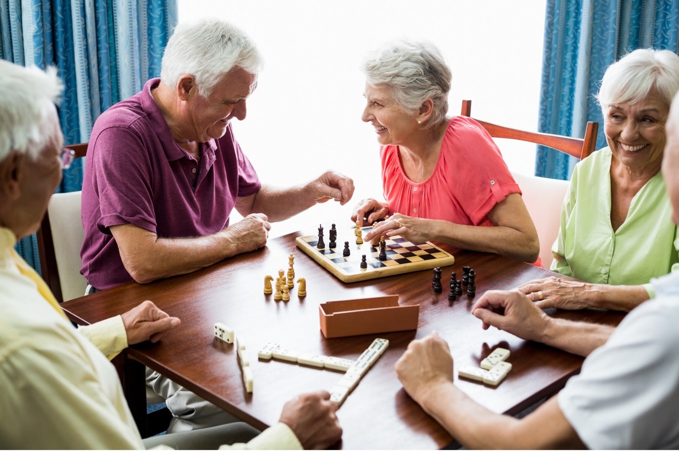 Group of elderly friends playing chess and dominoes at a table, smiling and enjoying each other's company.