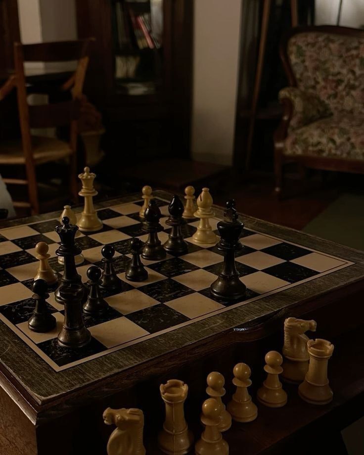 A dimly lit room with a chessboard in the foreground. Chess pieces are arranged on the board, and chairs are in the background.