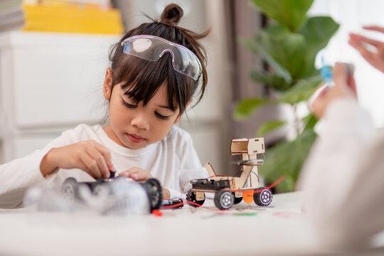 A girl wearing safety goggles is focused on assembling a toy car while another person is in the background, possibly helping.