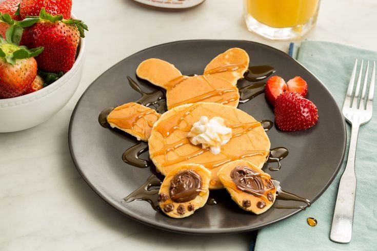 Pancakes shaped like a bunny with banana slices and chocolate, garnished with syrup and strawberries, beside a bowl of berries and juice.