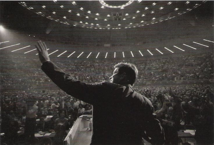 A person at a podium waves to a large audience in an arena with bright overhead lighting.