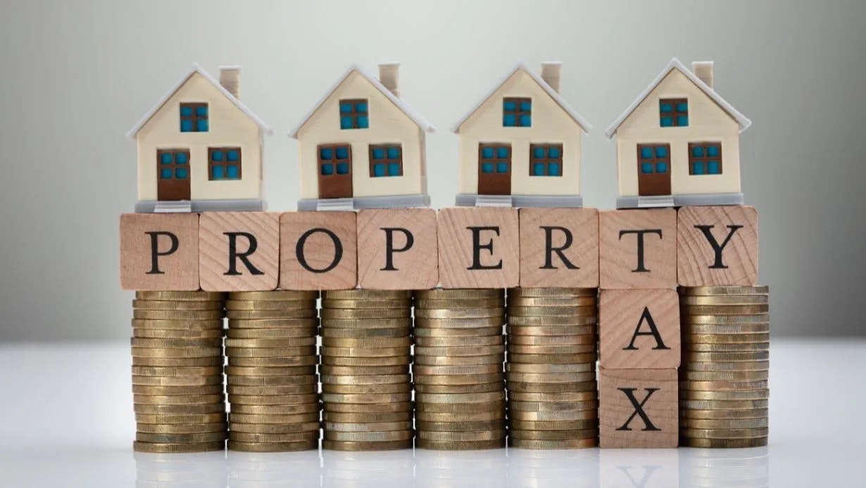 Miniature houses on stacked coins with blocks spelling "PROPERTY TAX," symbolizing property taxes and real estate value.