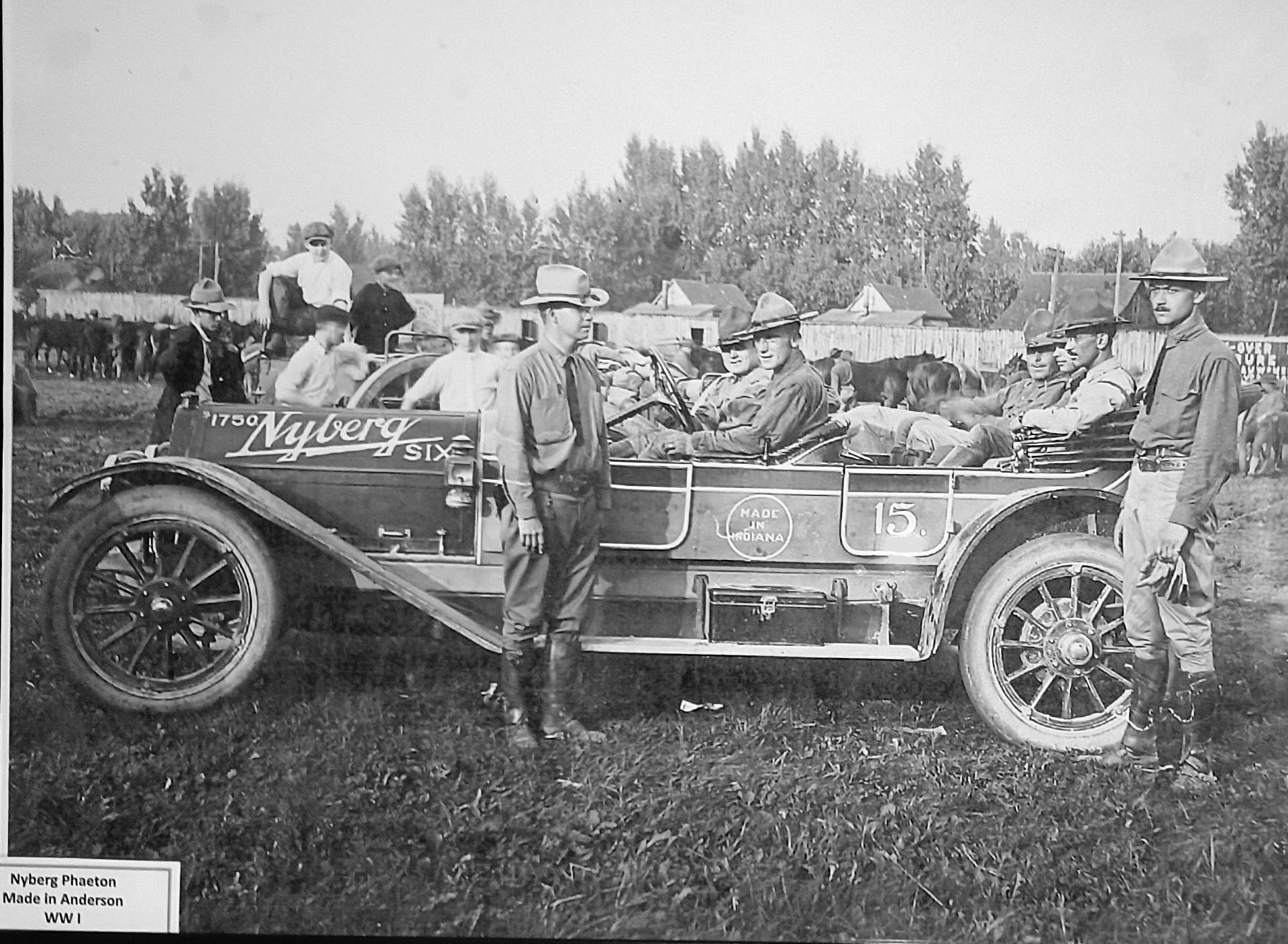 A vintage Nyberg Six car with soldiers seated inside, outdoors, with other people and a herd in the background.