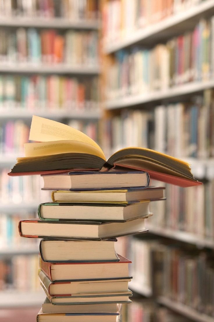 A stack of books with an open book on top, in front of blurred library shelves.