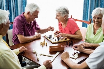 A group of elderly people playing chess and dominoes at a table, smiling and enjoying each other's company.