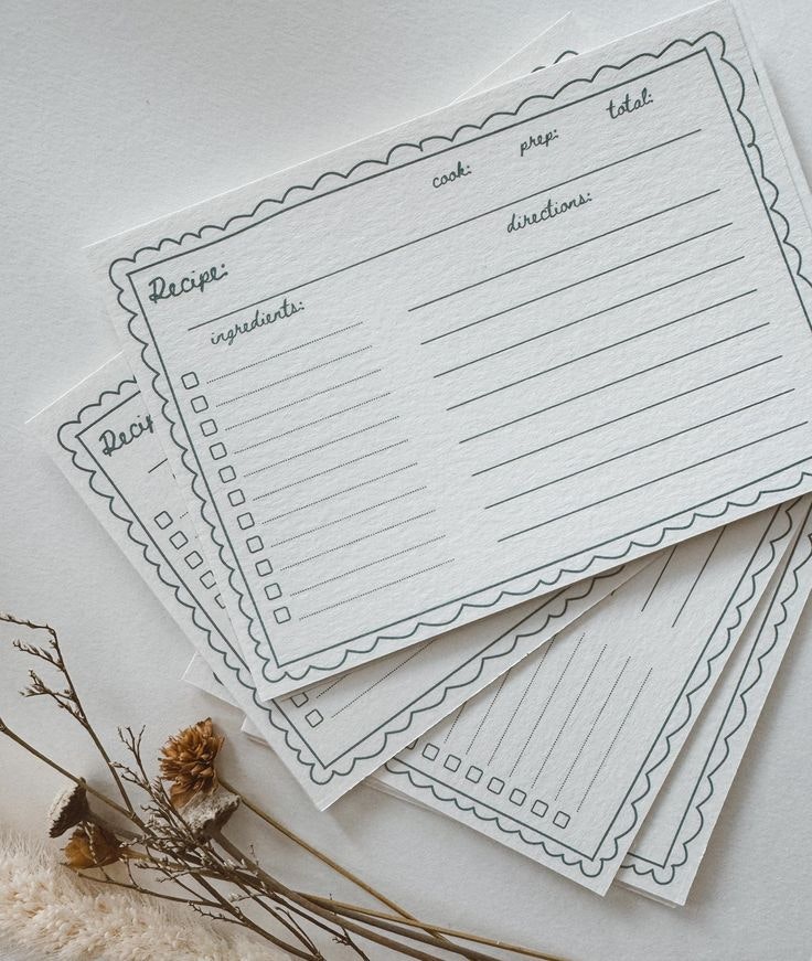 Blank recipe cards with sections for ingredients, directions, and cooking times, placed on a surface with dried flowers nearby.