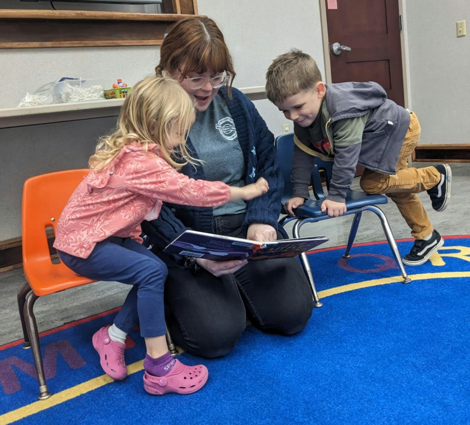 An adult reading to two children in a classroom, seated on bright-colored chairs on a blue carpet.