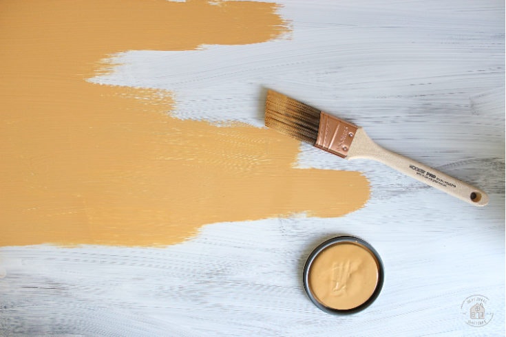 A paintbrush rests on a surface with a mustard-yellow stripe of paint next to a tin of similar colored paint.