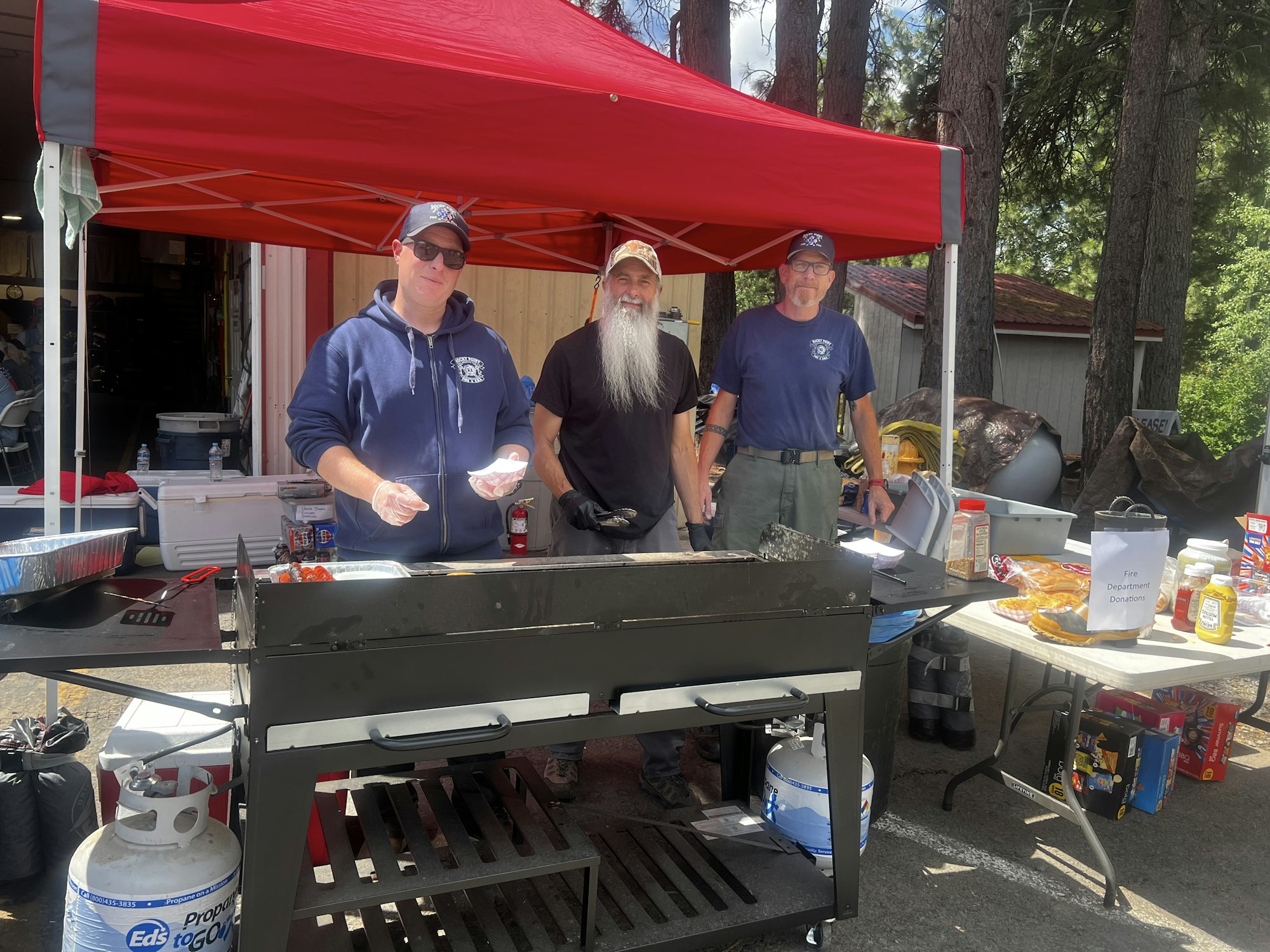 Three people at an outdoor grill with condiments and a donation box under a red canopy.