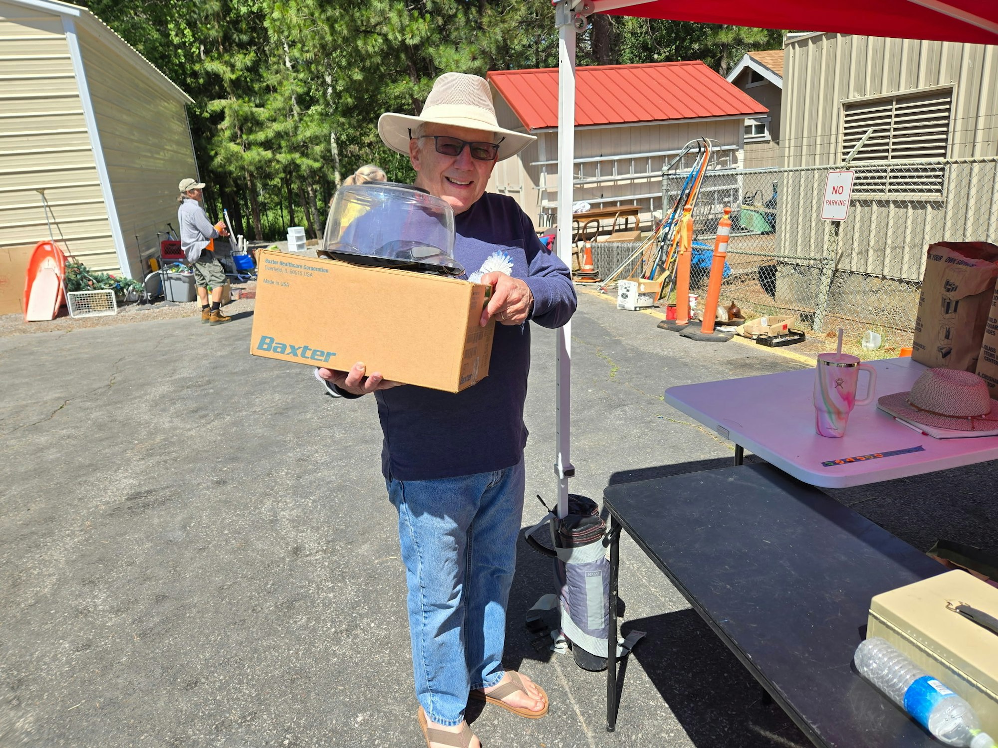 A person holds a box labeled "Baxter" under a red tent, with more items and a "No Parking" sign in the background.