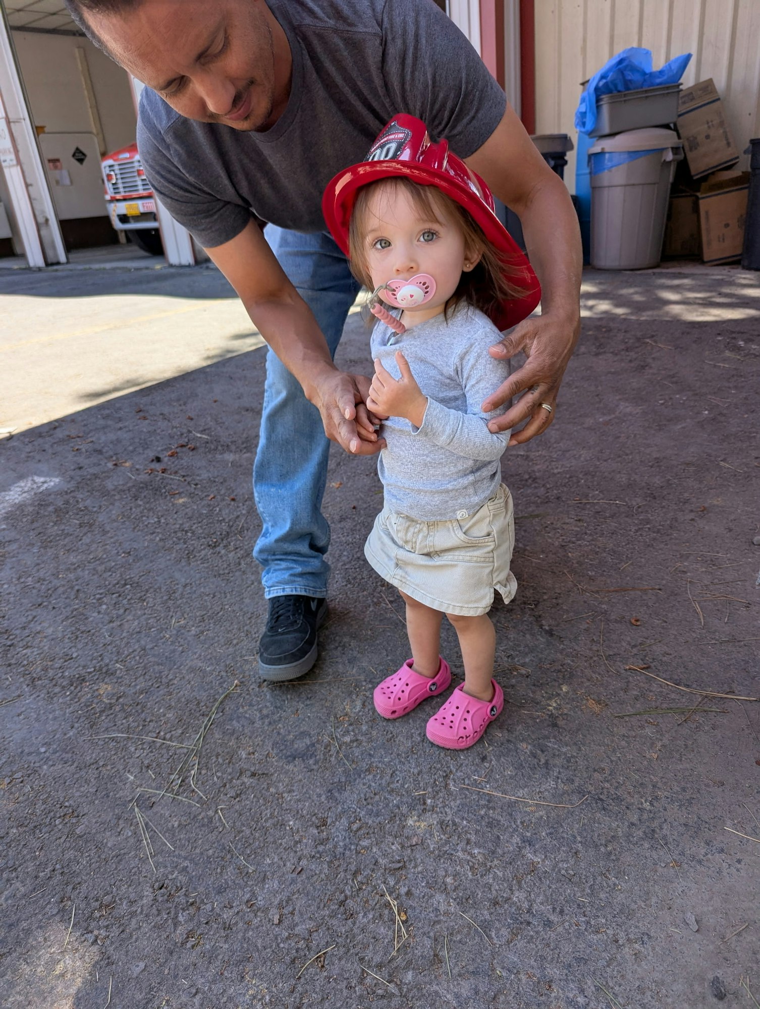 A man is helping a child wearing a red fire hat and pacifier, in an outdoor area with a few boxes and bins in the background.