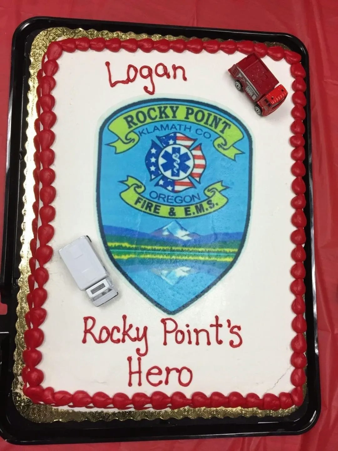 A cake with "Logan, Rocky Point's Hero" text, featuring a Rocky Point Fire & E.M.S. logo and toy fire trucks on top.