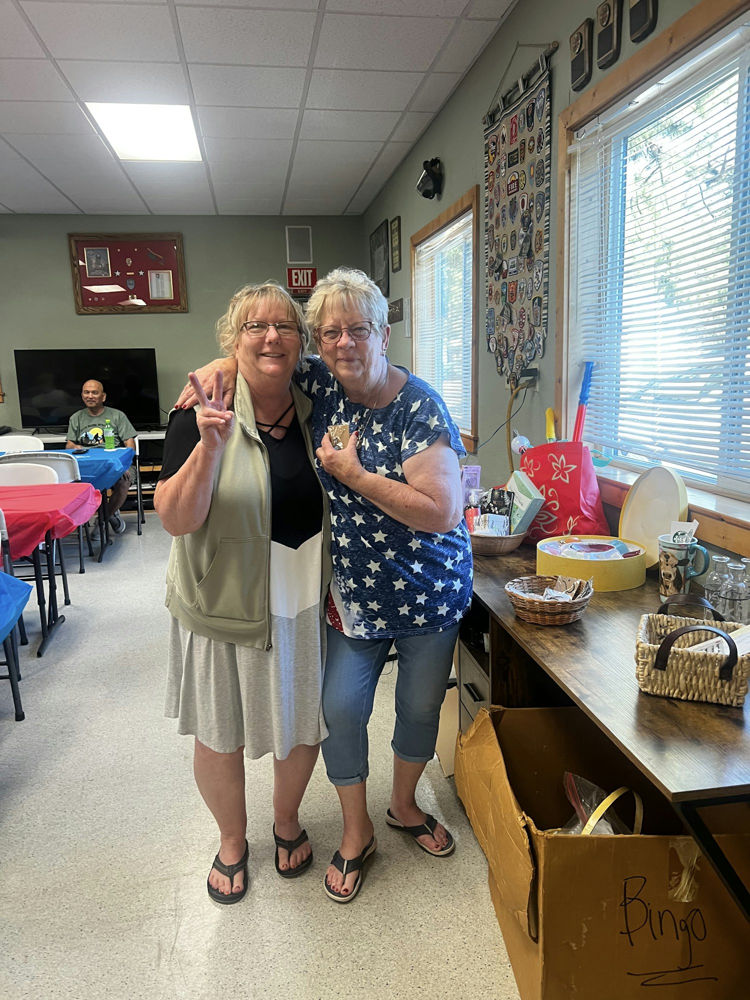 Two women posing together in a room with various items on a table and a "Bingo" box nearby.