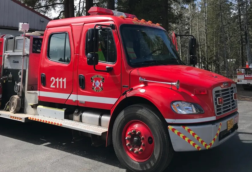 Red fire truck with number 2111, Rocky Point logo, parked outdoors near trees and a building.
