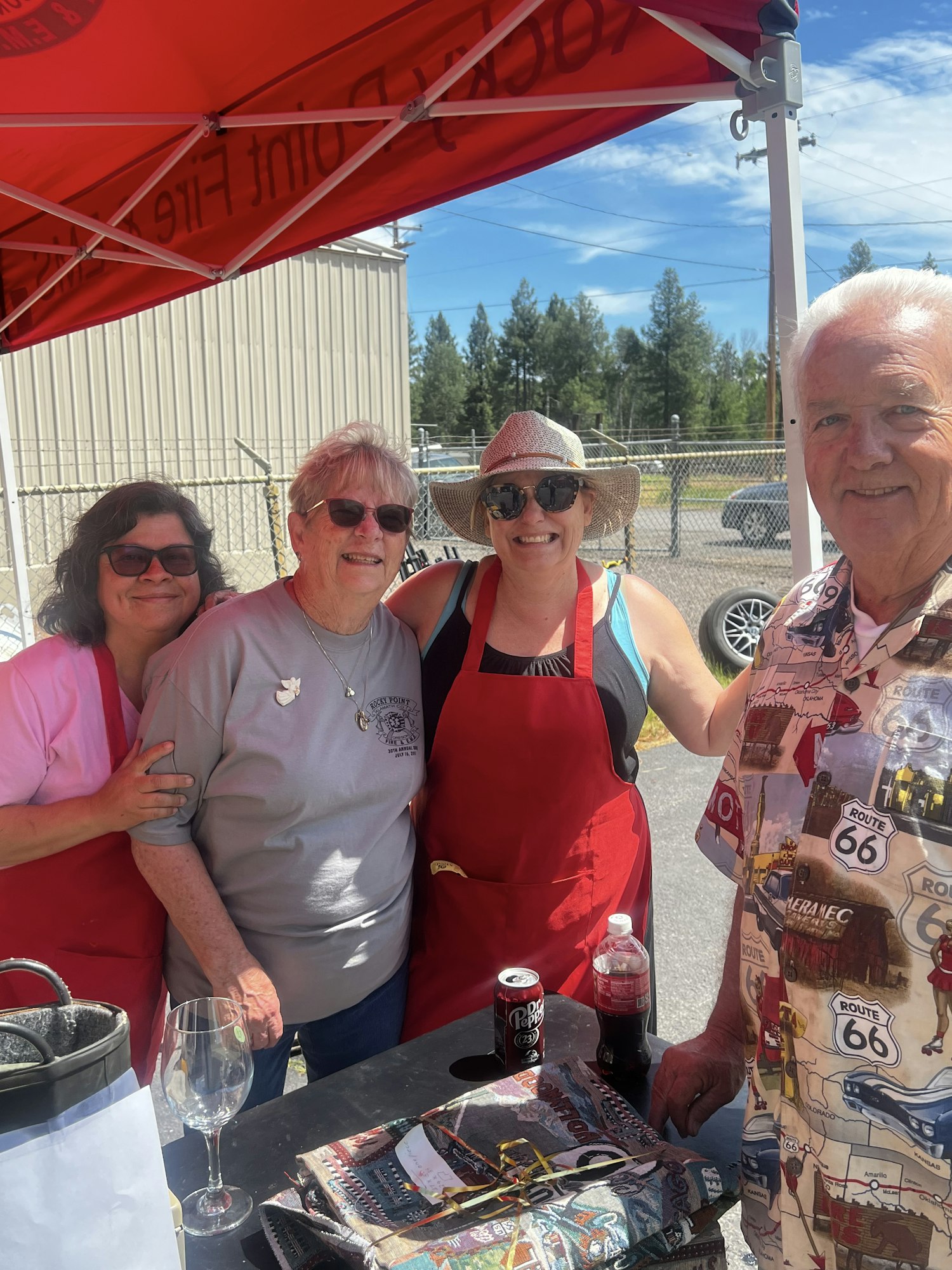 A group of four people smiling under a red canopy. One wears a Route 66 shirt, another has a sunhat. Wine and soda on a table nearby.