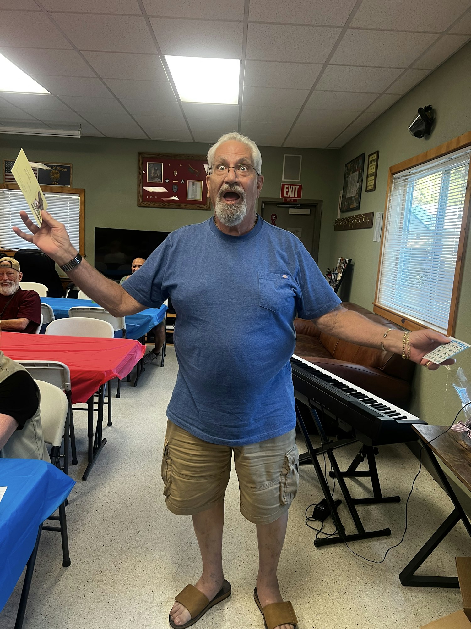 A man in a blue shirt and shorts, holding papers, stands next to a keyboard in a room with tables and chairs.