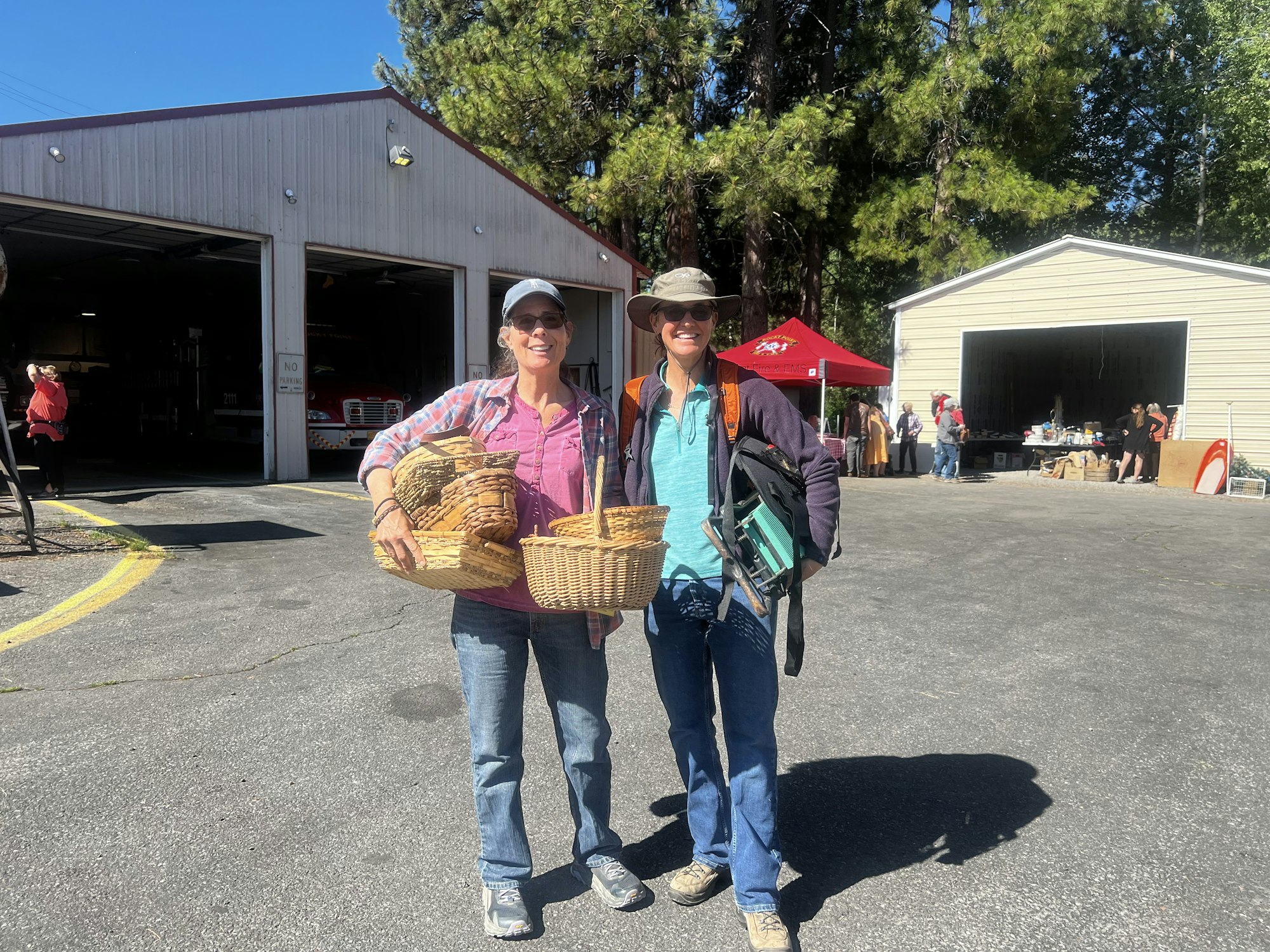 Two people standing outside holding baskets and chairs, with a building and a garage sale in the background.