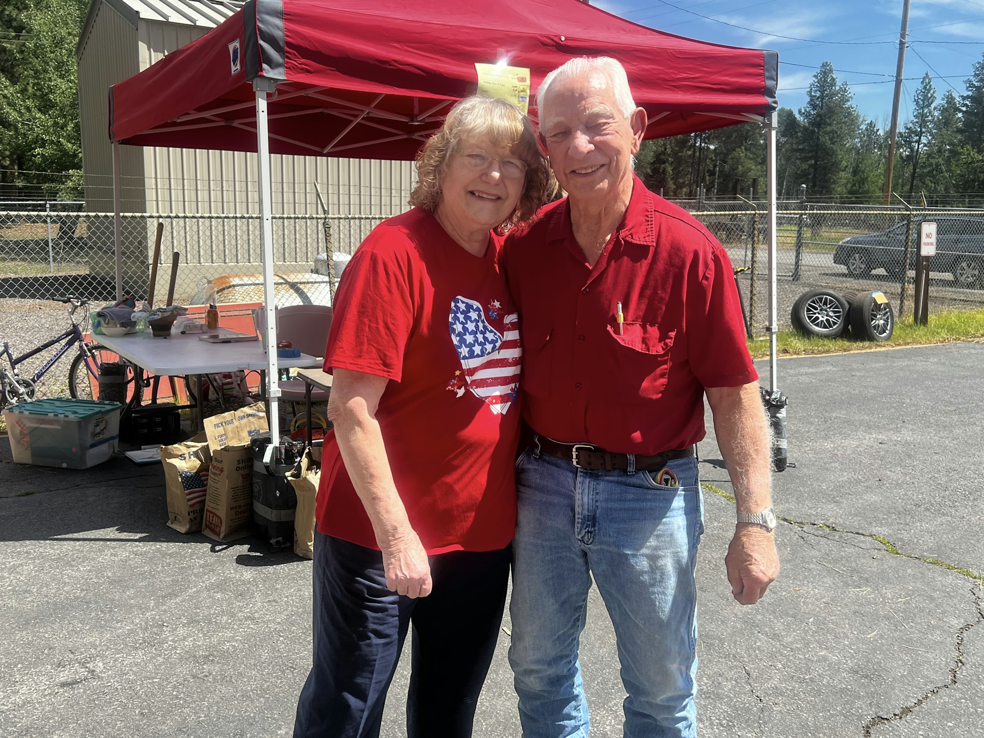 An older couple in red shirts posing together outside under a red canopy, near a table and various items.