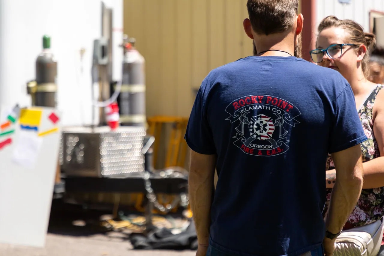 A person in a Rocky Point Fire & EMS shirt facing a woman, with tanks and equipment in the background.