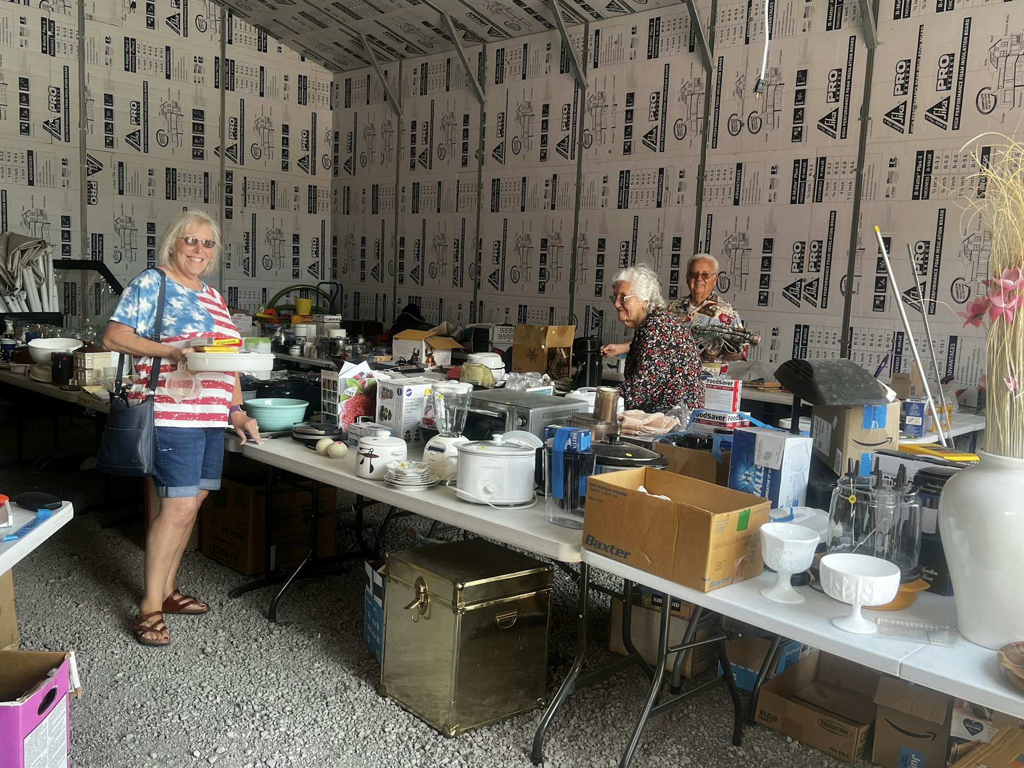 Three people in a garage-like space with tables full of household items, appearing to be at a sale or market.