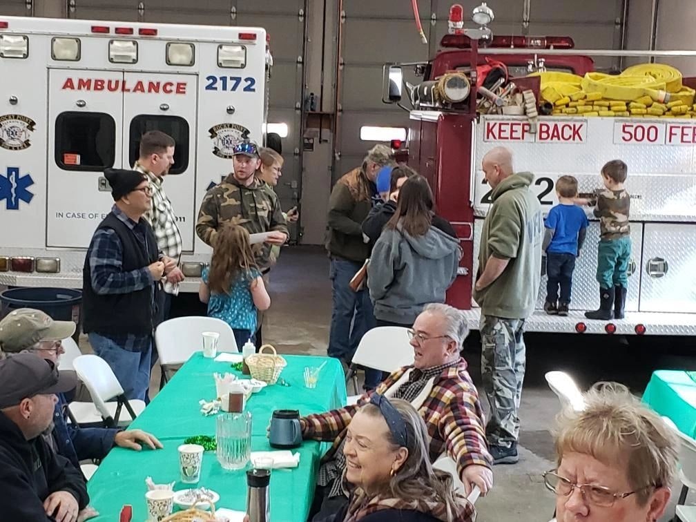 People gathered in a garage with an ambulance and fire truck, sitting at tables covered in green tablecloths, in casual clothing.