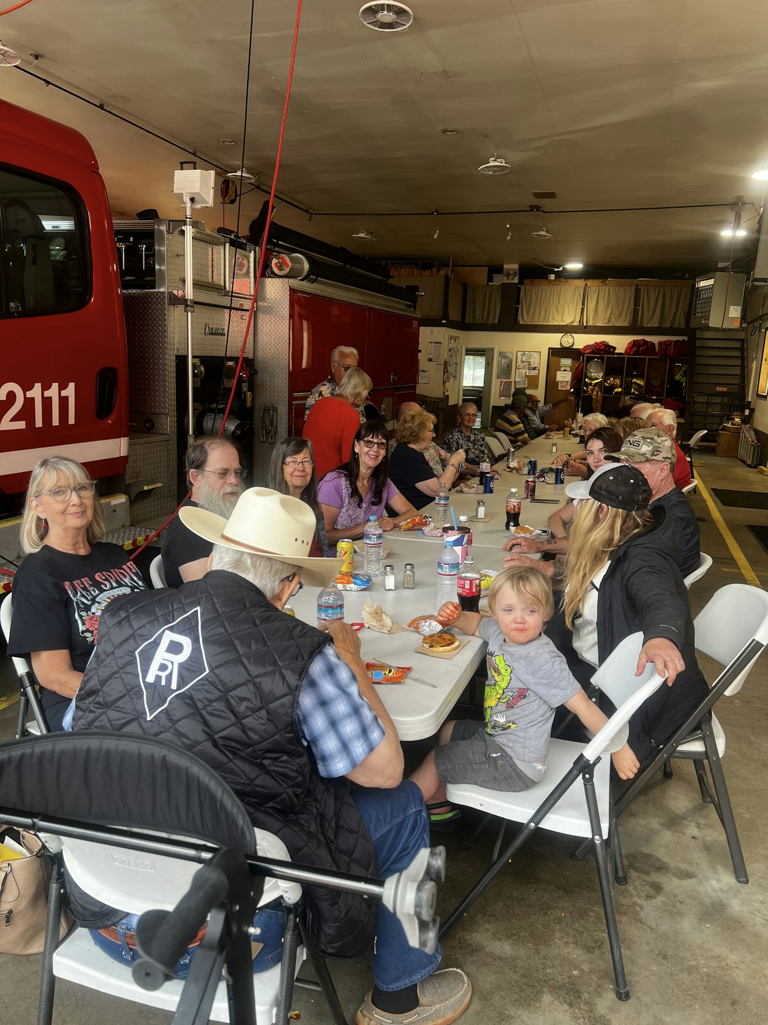 A group of people sitting at tables inside a garage, eating and socializing near a red vehicle.
