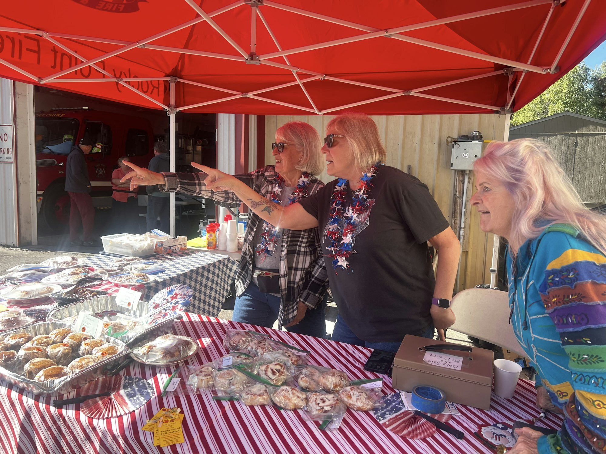 Three women pointing at something under a red tent, with baked goods on a table covered in striped cloth, and a firetruck in the background.