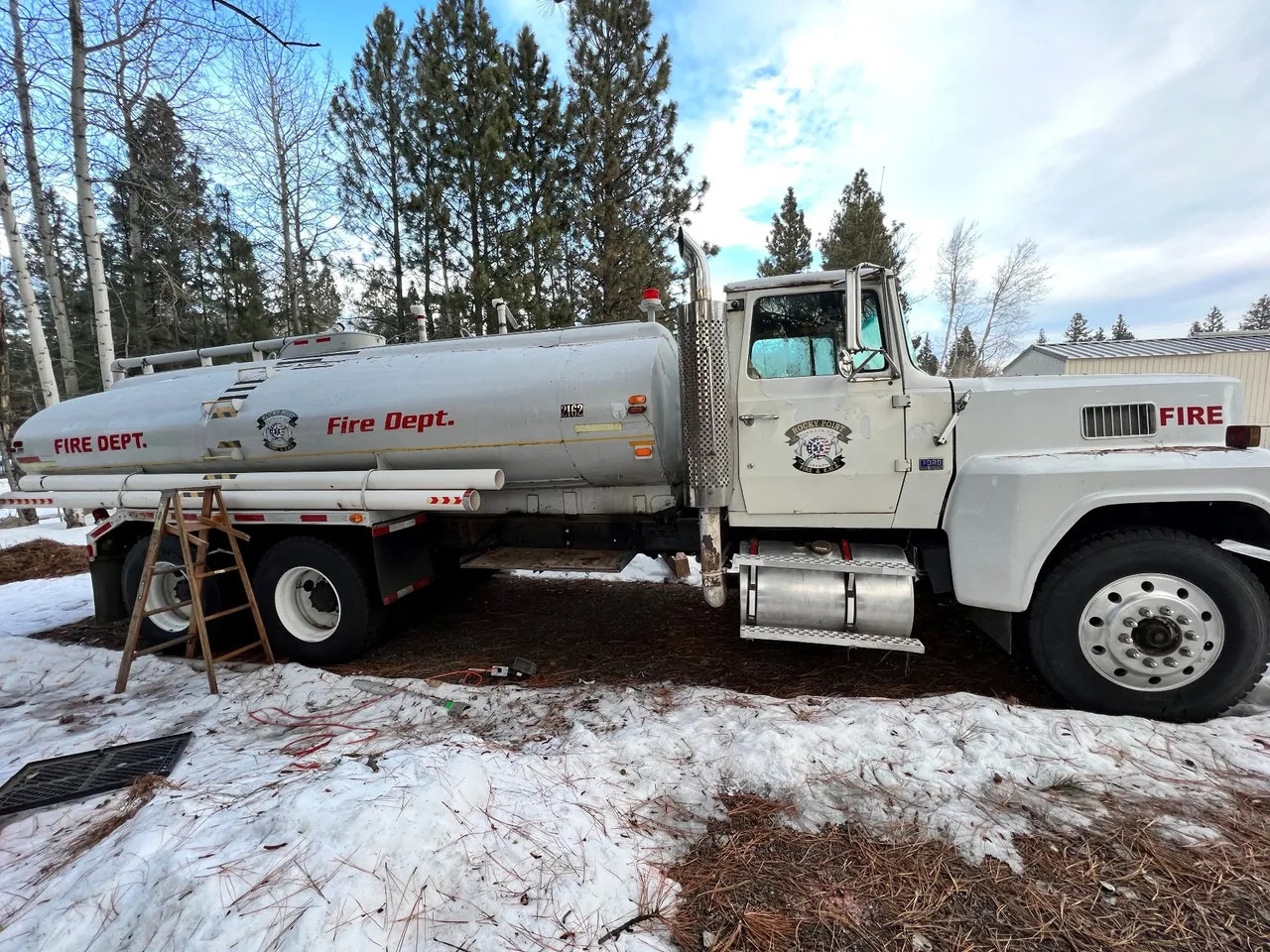 Fire department tanker truck in a snowy forested area with a ladder leaning against it.